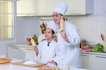 Millennial Asian young little cute boy pastry chef and male cooking teacher in uniform with white tall cook hat standing smiling holding wooden bakery rolling pin posing together in home kitchen