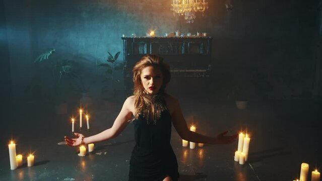 Fantasy halloween woman modern witch sits on knees in circle of burning candles dark room. Lady sorceress dances ritual dance, hands raised to night sky. Dark gothic queen girl in sexy black dress. 4k
