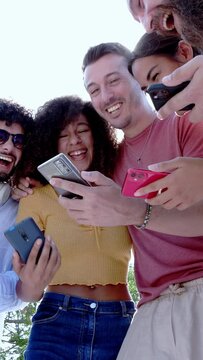 Vertical shot of young group of happy people smiling while using mobile phone together. Multiracial friends standing outdoors in the park holding cellphone. Technology, youth and social media concept