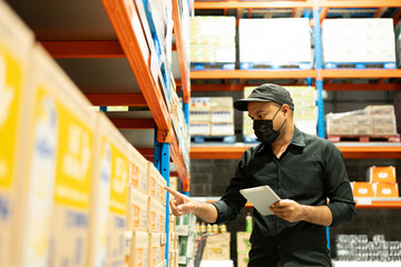 Warehouse worker wearing a hat and black shirt hands holding tablet check stock on tall shelves in warehouse storage. Asian auditor or staff work looking up stocktaking inventory in warehouse store.