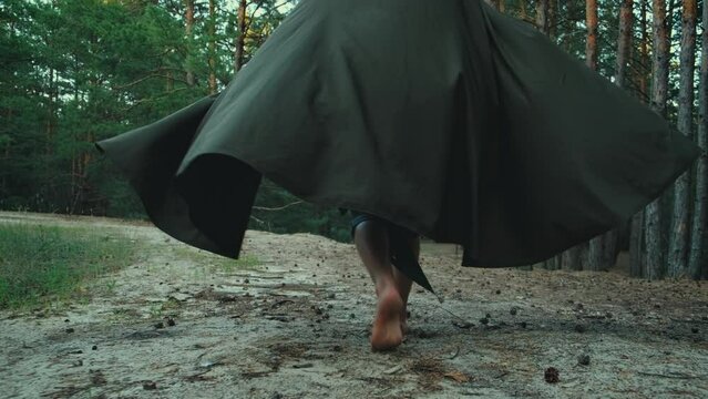 Fantasy young male elf prince walks in dark summer forest foot steps close up. guy Wizard man robber. Creative clothes cloak cape green costume waving in motion. Fashion model back rear view video 4k