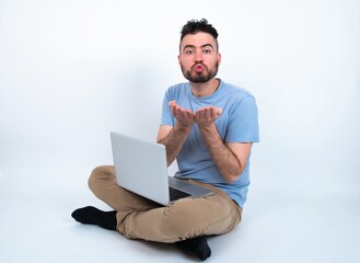 Young caucasian man with laptop sitting over white studio sending blow kiss with pout lips and holding palms to send air kiss.