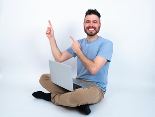 Young caucasian man with laptop sitting over white studio indicating finger empty space showing best low prices, looking at the camera