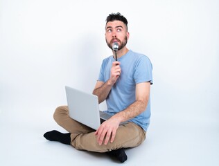 Very hungry Young caucasian man with laptop sitting over white studio holding spoon into mouth dream of tasty meal