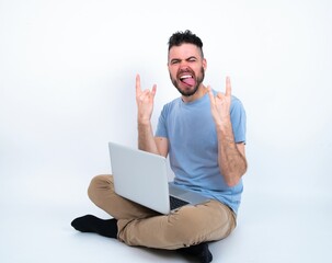 Young caucasian man with laptop sitting over white studio making rock hand gesture and showing tongue