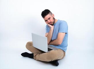 Very bored Young caucasian man with laptop sitting over white studio holding hand on cheek while support it with another crossed hand, looking tired and sick,