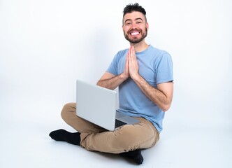 Young caucasian man with laptop sitting over white studio praying with hands together asking for forgiveness smiling confident.