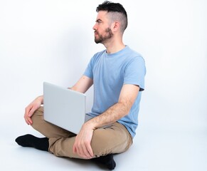 Young caucasian man with laptop sitting over white studio looking to side, relax profile pose with natural face with confident smile.