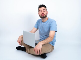 Shot of pleasant looking Young caucasian man with laptop sitting over white studio , pouts lips, looks at camera, Human facial expressions