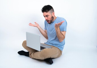 Frustrated Young caucasian man with laptop sitting over white studio feels puzzled and hesitant, shrugs shoulders in bewilderment, keeps mouth widely opened, doesn't know what to do.