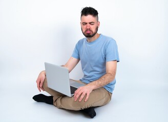 Dismal gloomy rejected Young caucasian man with laptop sitting over white studio has problems and difficulties, curves lower lip and closes eyes in despair, being in depression