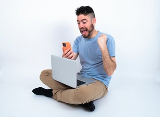 Positive Young caucasian man with laptop sitting over white studio holds modern cell phone connected to headphones, clenches fist from good emotions, exclaims with joy,