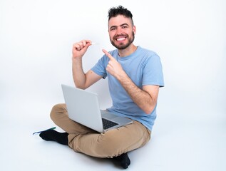 Young caucasian man with laptop sitting over white studio holding an invisible aligner and pointing at it. Dental healthcare and confidence concept.