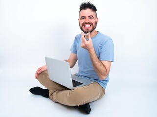 Young caucasian man with laptop sitting over white studio holding an invisible aligner ready to use it. Dental healthcare and confidence concept.