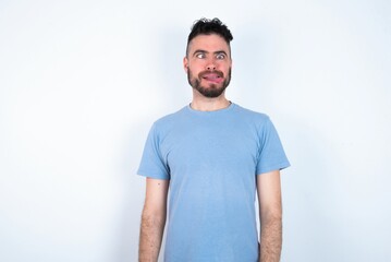 Fototapeta premium Funny Young caucasian man wearing blue T-shirt over white background makes grimace and crosses eyes plays fool has fun alone sticks out tongue.
