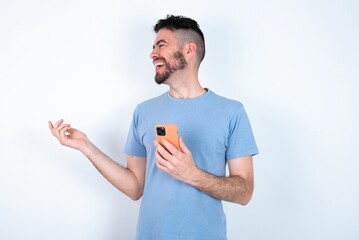 Happy pleased Young caucasian man wearing blue T-shirt over white background raises palm and holds cellphone uses high speed internet for text messaging or video calls