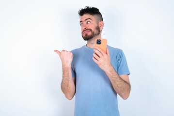 Young caucasian man wearing blue T-shirt over white background points thumb away and shows blank space aside, holds mobile phone for sending text messages.