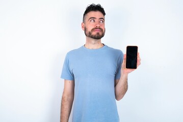 Young caucasian man wearing blue T-shirt over white background holds new mobile phone and looks mysterious aside shows blank display of modern cellular