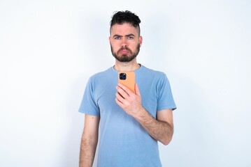 Upset dissatisfied Young caucasian man wearing blue T-shirt over white background uses mobile software application and surfs information in internet, holds modern mobile hand