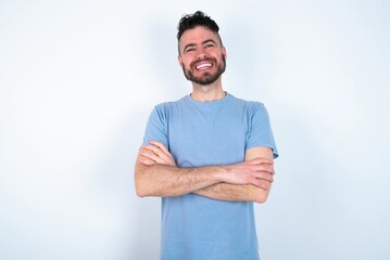 Portrait of charming Young caucasian man wearing blue T-shirt over white background standing confidently smiling toothily with hands folded