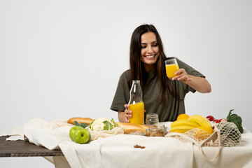 Woman in green t-shirt pouring a juice from a glass bottle in a glass over a table with mesh eco bag, healthy vegan vegetables, fruits, bread, snacks. Zero waste concept.