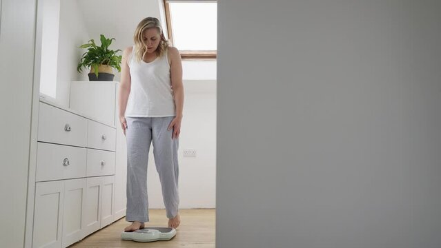 Menopausal Mature Woman Concerned With Weight Gain Standing On Scales In Bedroom At Home