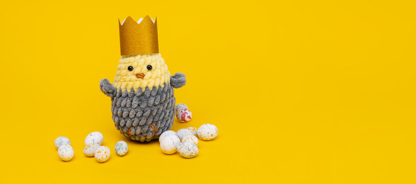 Knitted soft chick in a golden crown with easter covered chocolate eggs on a yellow background with copy space. Easter banner
