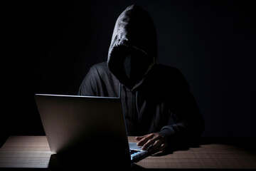 Hacker wearing black hoodie using laptop computer while working on dark background. Created with Generative AI Technology