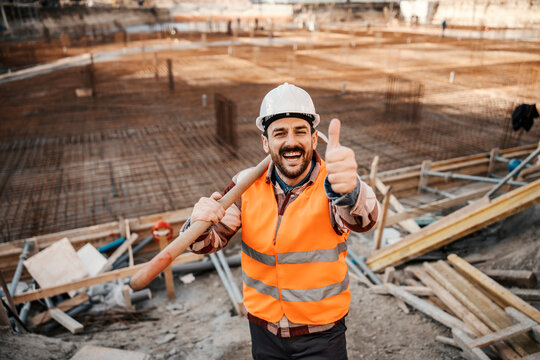A builder is holding shovel on a shoulder and giving thumbs up while standing on site and smiling at the camera.