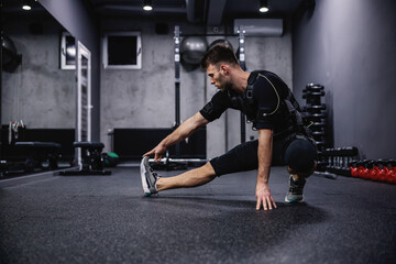 Fototapeta na wymiar A young sportsman in a special EMS suit stretches his legs and warms up his muscles for training indoor gym. Fitness in EMS suit in a modern gym concept, revolutionary training, electrical stimulation