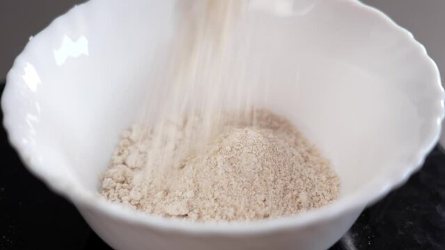 Pouring oat flour into a white ceramic bowl for cooking dessert in slow motion close up