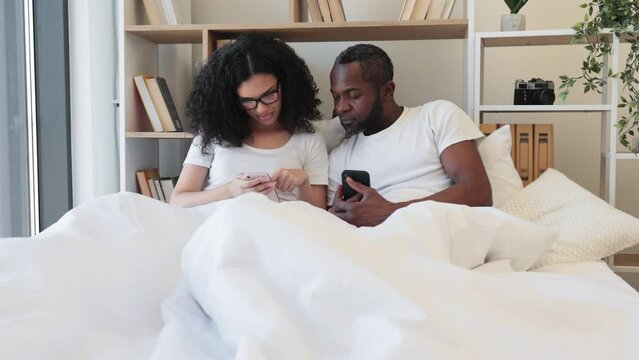 Smiling multicultural husband and wife surfing internet using smartphones while sitting under cozy duvet in bedroom of modern flat. Busy spouses networking through social media via digital devices.