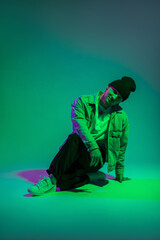 Handsome happy professional dancer man in fashionable denim clothes sits in the studio on a colored background with green and purple lights