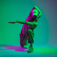 Stylish hip hop dancer professional with a cap in fashionable clothes with sneakers dances in the studio with green and purple light