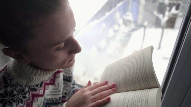 Portrait of happy young woman in knitted Christmas sweater reading favorite book by the window at home in winter time
