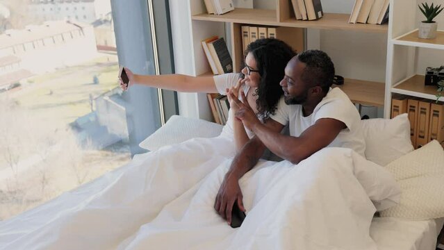 Smiling intercultural woman in glasses taking selfie of her and her husband showing peace gesture while relaxing in bed at home. Married couple shooting photos on mobile for sharing in social media.