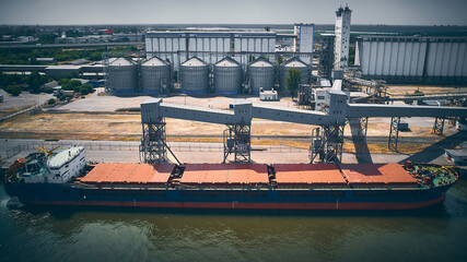 Bulk carrier ship in cargo port. Aerial view of barge in a dock. Grain elevator and granary silos...