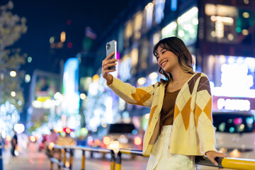 Young Asian woman using mobile phone taking selfie or video call in the city at night. Attractive girl enjoy and fun modern outdoor nightlife  travel city street with using portable device technology.