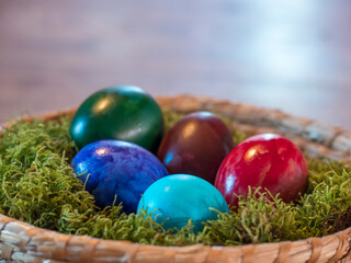 Various coloured decorated easter eggs in wicker basket nest with moss and colored Easter eggs on beige wooden bokeh background. For Happy Easter holiday card. Selective focus, copy space