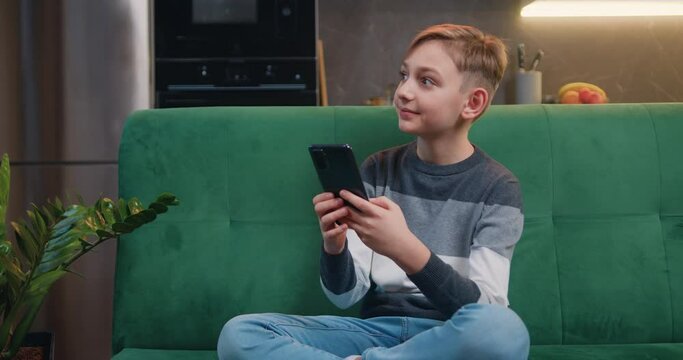 Beautiful caucasian boy of 9 years old sitting on sofa and playing mobile game on smartphone at home. Teenager playing mobile phone. Kid using phone for gaming.