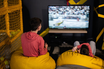 Two brothers playing football video game console, sitting on yellow pouf in kids play center.