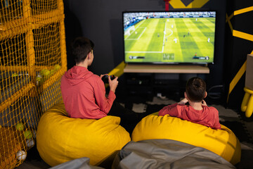 Two brothers playing football video game console, sitting on yellow pouf in kids play center.