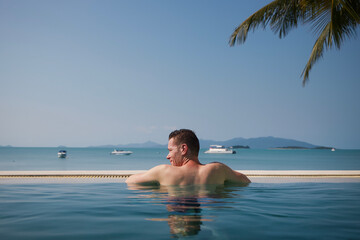Rear view man while relaxing in swimming pool against sea with islands. Vacation on beach in tropical destination..