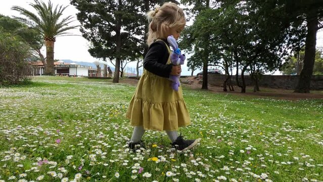 Little girl with a plush hare walks through the park with blooming daisies