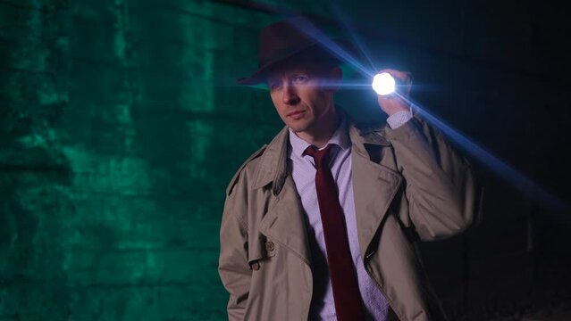stylish portrait of a searching detective in a raincoat and hat with a flashlight looking for clues or retro style 1950 slow mo