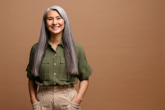Cheerful smiling stylish confident 50 years old Asian beautiful woman standing looking at camera at brown background. Grey haired middle-aged woman isolated, copy space