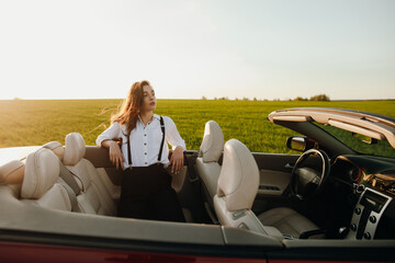 young beautiful woman in a white shirt sitting in a red car cabriolet with a white interior. red car in the field on a sunny day. the wind blows the girl's hair. red cabriolet.