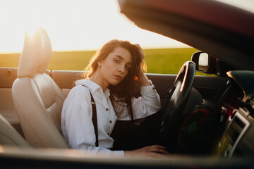 Fototapeta na wymiar young beautiful woman in a white shirt sitting in a red car cabriolet with a white interior. red car in the field on a sunny day. the wind blows the girl's hair. red cabriolet.