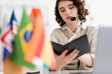 young brunette language teacher in headset writing in notebook near international flags on blurred foreground. © LIGHTFIELD STUDIOS
