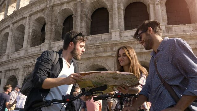 Three happy young friends tourists with bikes and backpacks at Colosseum in Rome reading map guide for directions on sunny day slow motion steadycam ground shot